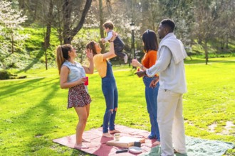 Cheerful multi ethnic family enjoying a sunny picnic on a green lawn in the park, happily lifting