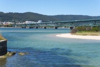 A long bridge crosses a shimmering blue river, surrounded by hills and a town in the background,