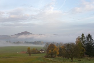 Fog in the valley, surrounded by trees and a small house, with a range of hills in the background,