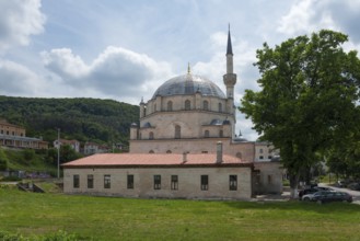 Mosque with dome and minaret, in front of it a car park with cars and a hill in the background,