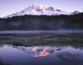 Snow capped mountain reflecting in a calm lake at dusk, surrounded by mist and lush forest. The sun