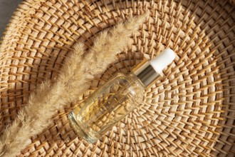 A glass cosmetic with golden essential oil rests on a woven basket alongside dried grass. The