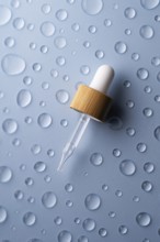 A glass pipette bottle lies on a textured blue surface Water droplets surround the bottle,