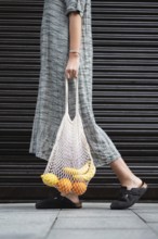 A woman in a long, gray dress carries a reusable mesh bag filled with bananas and oranges. The