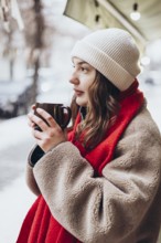 A young thoughtful woman in a white beanie and red scarf holds a warm mug, gazing thoughtfully