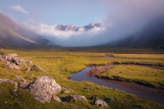A serene mountain valley landscape with a winding river and mist shrouded peaks in summer. The warm