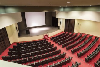 An empty convention center featuring rows of seats facing a wooden stage and a large white screen,