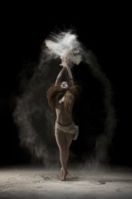 Slim young barefoot woman throwing sand over head while dancing against black background