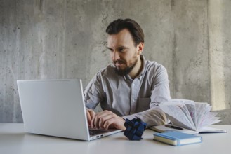 A man in a shirt works intently on a laptop at office. Beside him are a book and a Rubik cube,