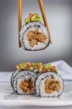 A close up of a sushi roll with vibrant avocado and salmon topping, held by chopsticks with more