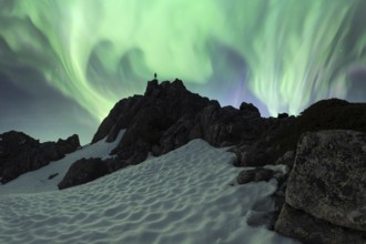A breathtaking Aurora Borealis illuminates the night sky above a snowy, rugged mountain terrain. An
