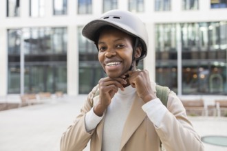 A black woman smiles while adjusting her helmet in an urban setting She is dressed modernly,
