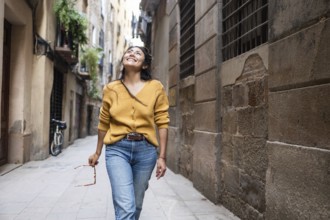 A Latin woman in a mustard sweater and jeans walks joyfully through a rustic urban alley, holding