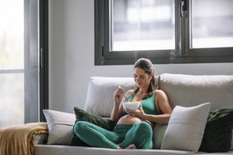 A pregnant woman sits comfortably on a couch in a yoga outfit, enjoying a relaxing moment while