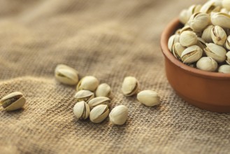 Close up of a clay bowl filled with pistachios on a burlap surface. Several pistachios are