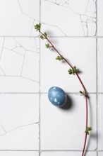 Top view of a single blue Easter egg adorned with a fresh green sprig on a textured white tiled