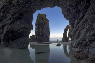 A person stands between towering sea arches, reflected in calm tidal pools, under a serene sky. The