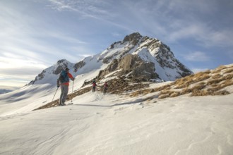 A group of ski tourers makes their way up a snow-covered mountain slope under a clear blue sky. The