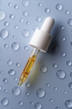 A close-up of a pipette filled with vitamin C serum on a blue surface decorated with water droplets