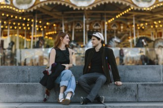 A young couple sits on stone steps, engaged in conversation, with a vibrant carousel in the