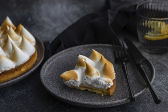 A slice of a homemade lemon cake sits elegantly on a dark plate, accompanied by a fork and knife. A