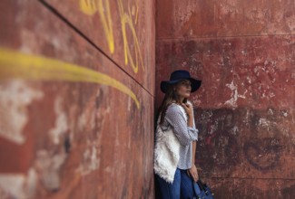 A fashionable woman in a blue hat leans against a weathered red wall adorned with graffiti. She