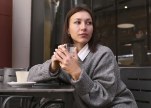 A thoughtful woman in a gray sweater holds a glass while sitting at an outdoor bar table. The cozy