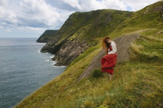A young woman wearing a flowing red dress and white blouse explores the rugged, green hillside