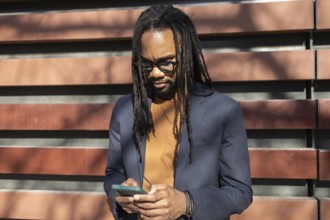 A cuban business professional with glasses and dreadlocks stands outdoors in sunlight, using a