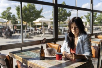 A woman sits alone in a sunlit cafe, looking down at her phone. Natural light flows through large