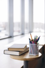 Books with dark covers stacked atop a circular wooden table beside a mesh pencil holder filled with