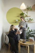 Two women engage in conversation in a cozy cafe setting. One sits with a tablet while the other