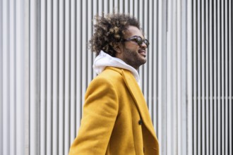 Afro man wearing a stylish mustard coat gazes confidently to the side. His sunglasses reflect a