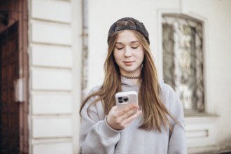 A teenage girl wearing a hoodie and cap is focused on her smartphone while standing outside. She is
