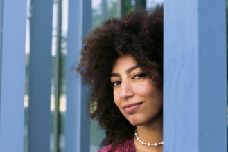 A young Arab woman with curly hair smiles confidently looking at camera, framed by blue columns.