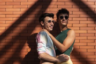 A happy gay couple in vibrant attire, embracing joyfully against a sunlit brick wall. They wear