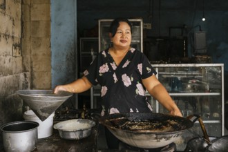 A woman stands in a rustic kitchen, preparing traditional dishes with a large wok. The worn walls