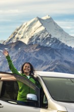 A woman stands joyfully in front of her car, surrounded by the breathtaking autumnal beauty of