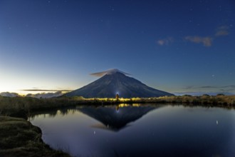 Back view of unrecognizable person gazing at Mount Taranaki's majestic peak reflected in a tranquil