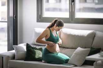 A pregnant woman, wearing a yoga outfit, sits on her knees on a couch, concentrating on well-being