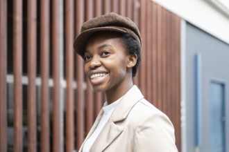 A confident black woman smiles warmly in an urban setting She wears a fashionable jacket and cap,