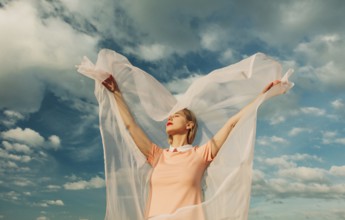 A woman with outstretched arms embraces the breeze, her sheer fabric flowing gracefully. A serene