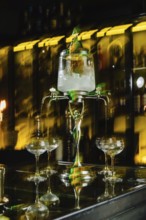 A vintage absinthe fountain pours into glasses on a dark bar counter. The ambiance is enhanced by a