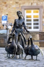 Bronze statue of a girl with geese on cobblestones, in the background a building with white