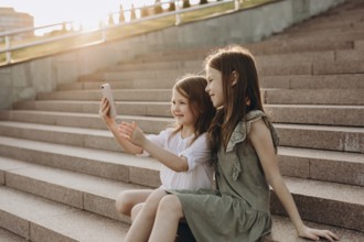 Two young girls sit on sunlit steps in summer, happily taking a selfie. They are enjoying a sunny