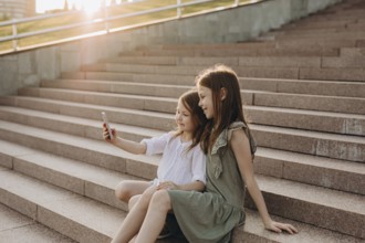 Two young girls sit together on sunlit stone stairs, smiling as they take a selfie. The warm glow