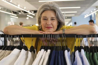 A mature woman with gray hair and a bright yellow shirt smiles warmly while leaning on a clothing