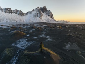 A man stands on a textured, frost-covered dune, gazing at the towering, snow-draped mountains of