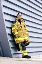 A young girl stands confidently against a modern wall while wearing a firefighter suit, showcasing
