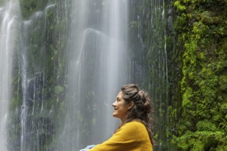 A woman in a yellow sweater enjoys a tranquil moment beside a lush green waterfall, surrounded by